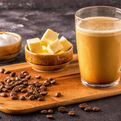 chopping board with a seethrough mug containing bulletproof coffee with coffe beans, coconut oil and butter on the side.